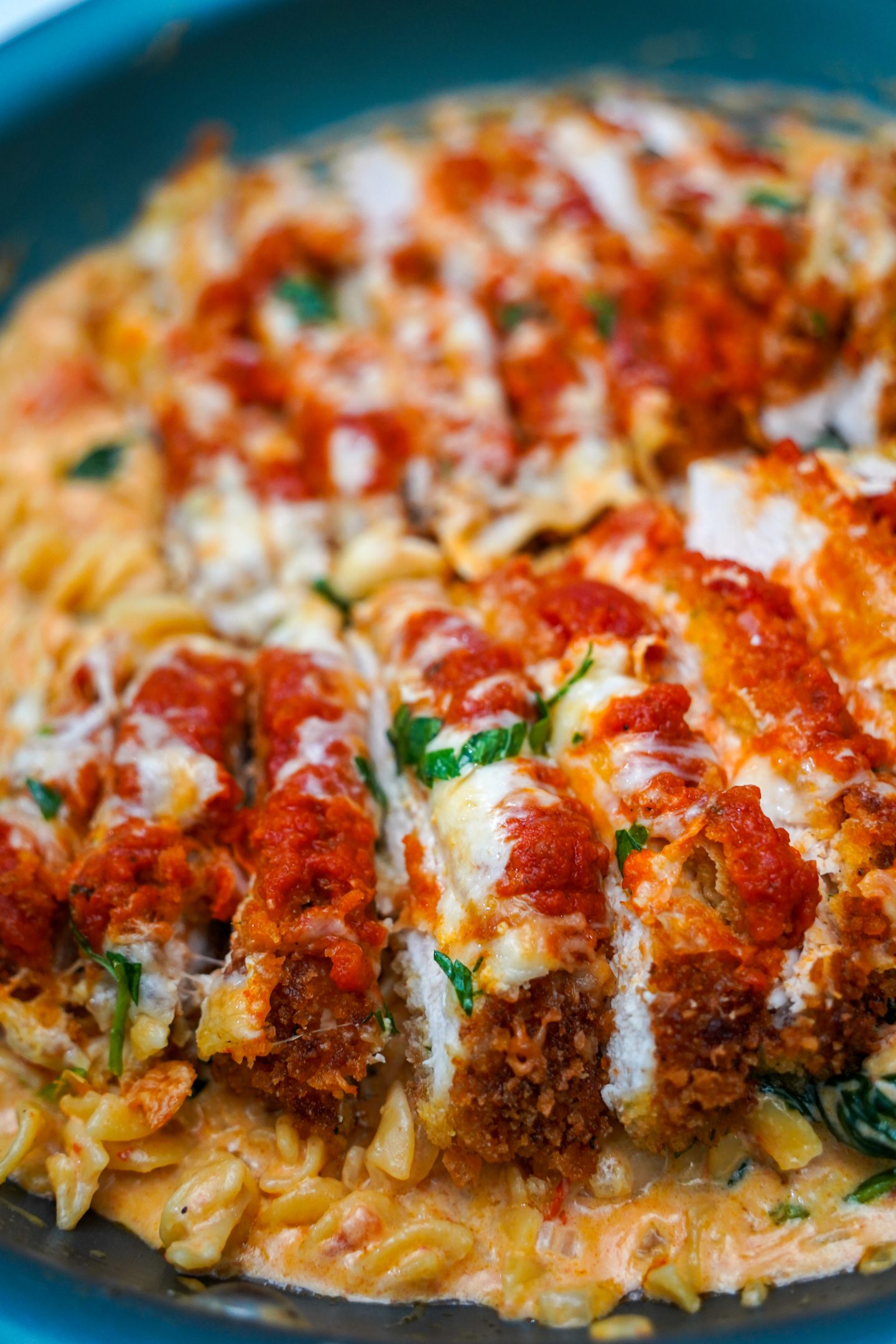 Close-up of crispy chicken parmesan sliced and served over creamy rotini pasta with spinach and cherry tomatoes in a rich, cheesy sauce, garnished with melted mozzarella and fresh herbs.