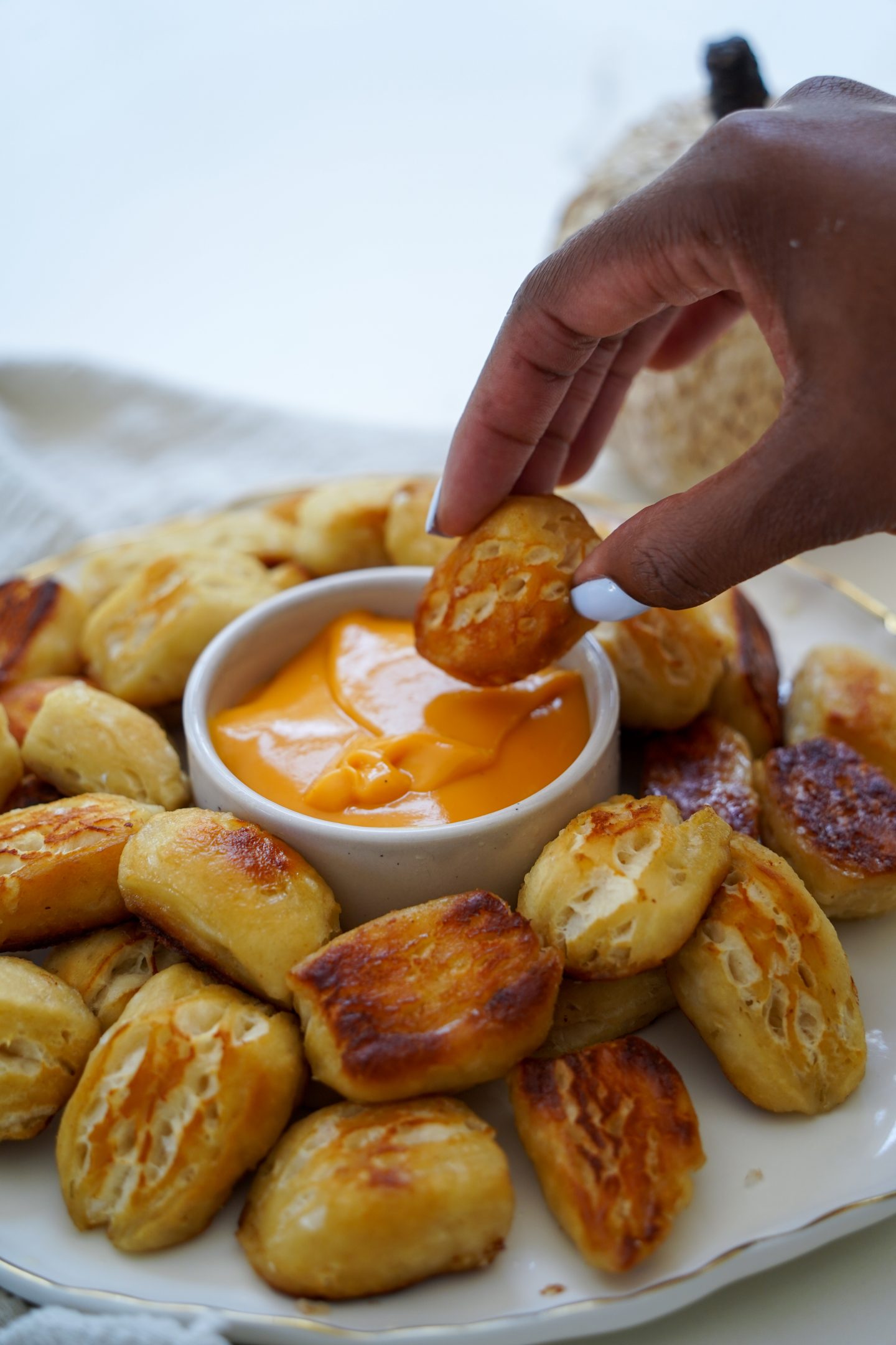 Golden-brown, bite-sized soft pretzel pieces arranged on a rustic wooden board. The pretzel bites are perfectly cooked with a shiny, salted exterior. A small bowl of mustard dipping sauce is visible in the corner, adding a pop of yellow color to the image. The background suggests a cozy kitchen setting, perfect for game day or fall snacking.