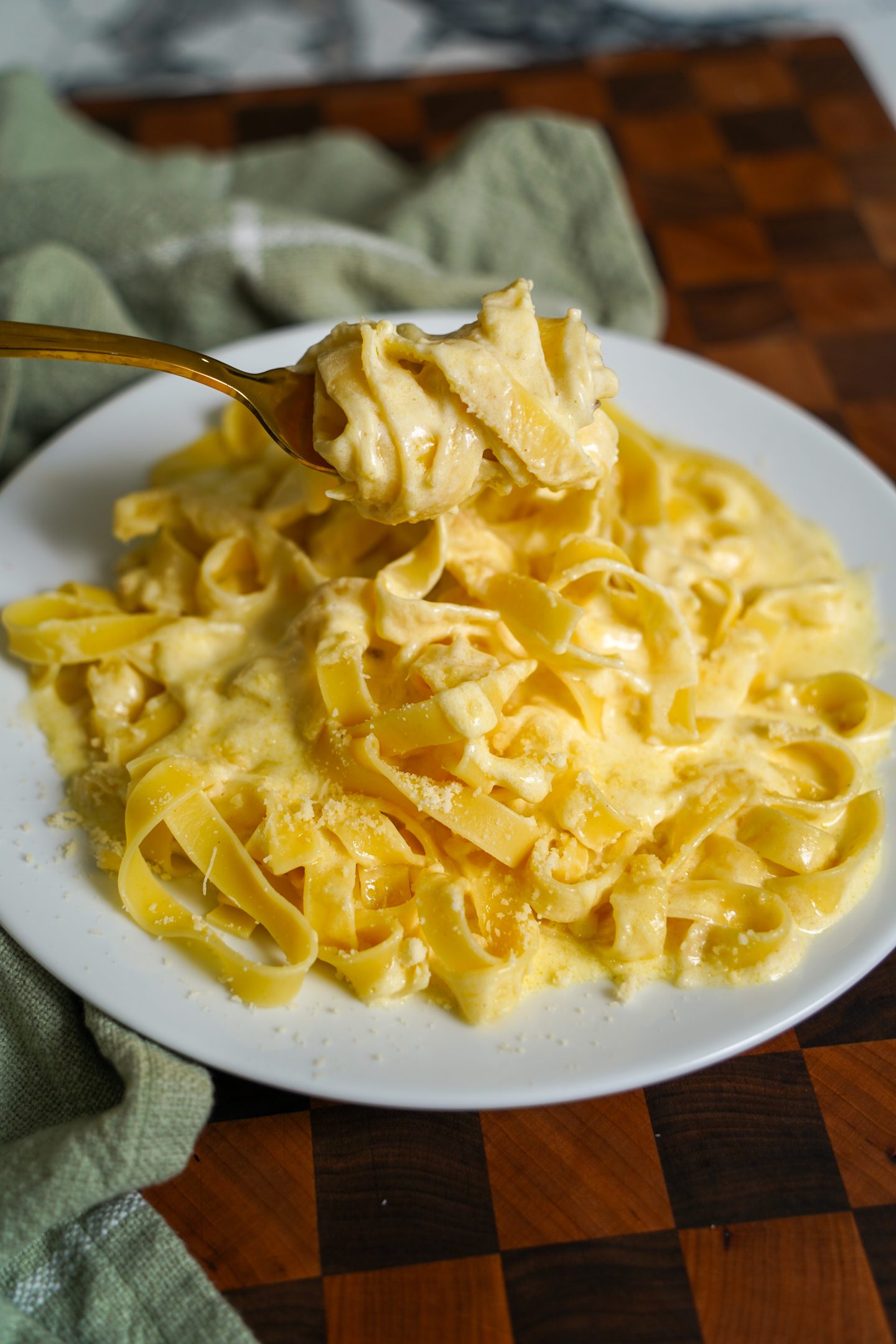 A close-up of a plate of creamy fettuccine alfredo with golden pasta coated in a rich, velvety sauce. A fork lifts a bite of the pasta, showcasing its smooth texture and generous Parmesan topping. The dish is served on a white plate over a wooden cutting board with a green linen napkin in the background.