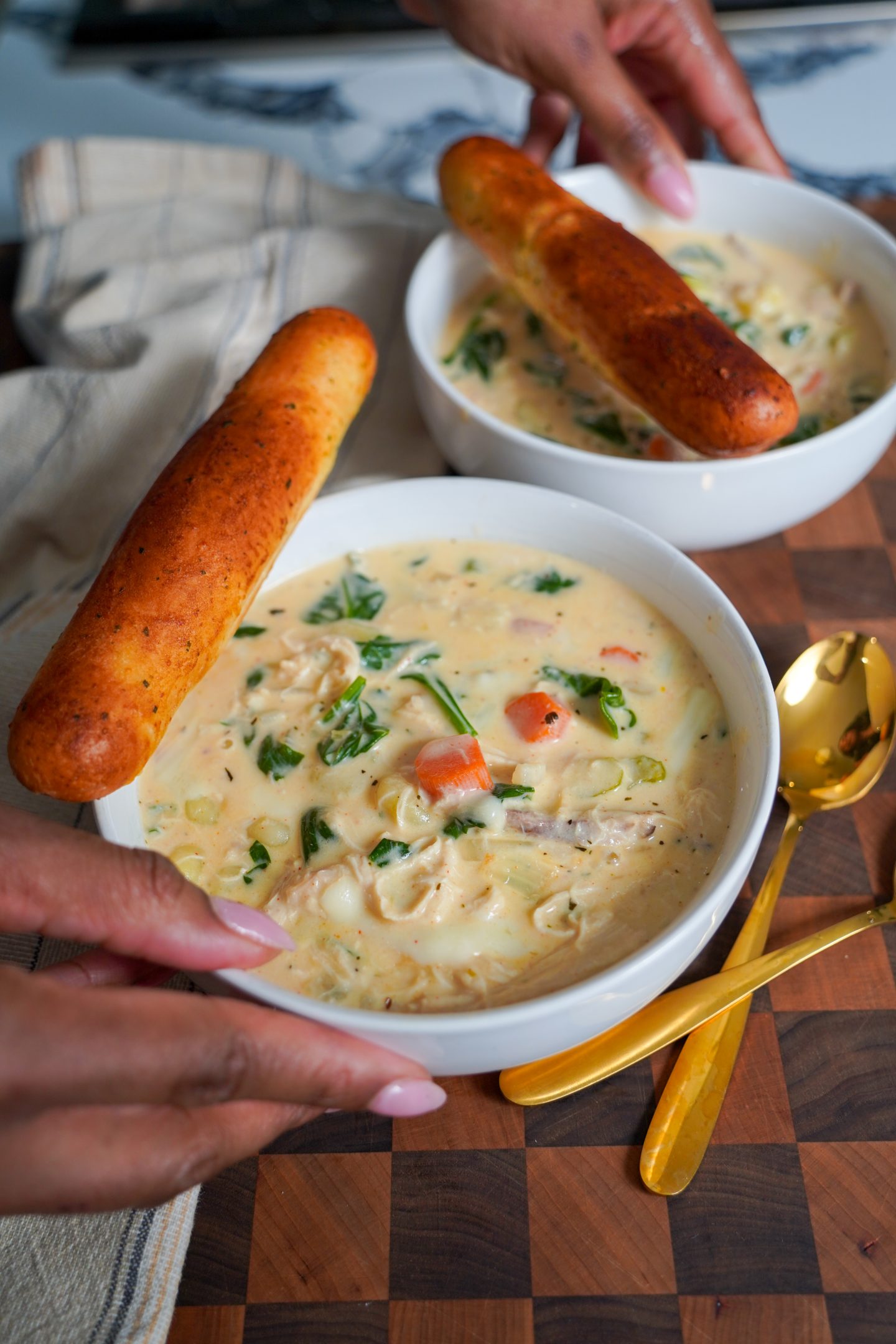 A close-up of two white bowls filled with creamy Chicken Gnocchi Soup, garnished with fresh spinach, carrots, and herbs, served with golden-brown breadsticks on a wooden board. A hand is reaching for one of the bowls.