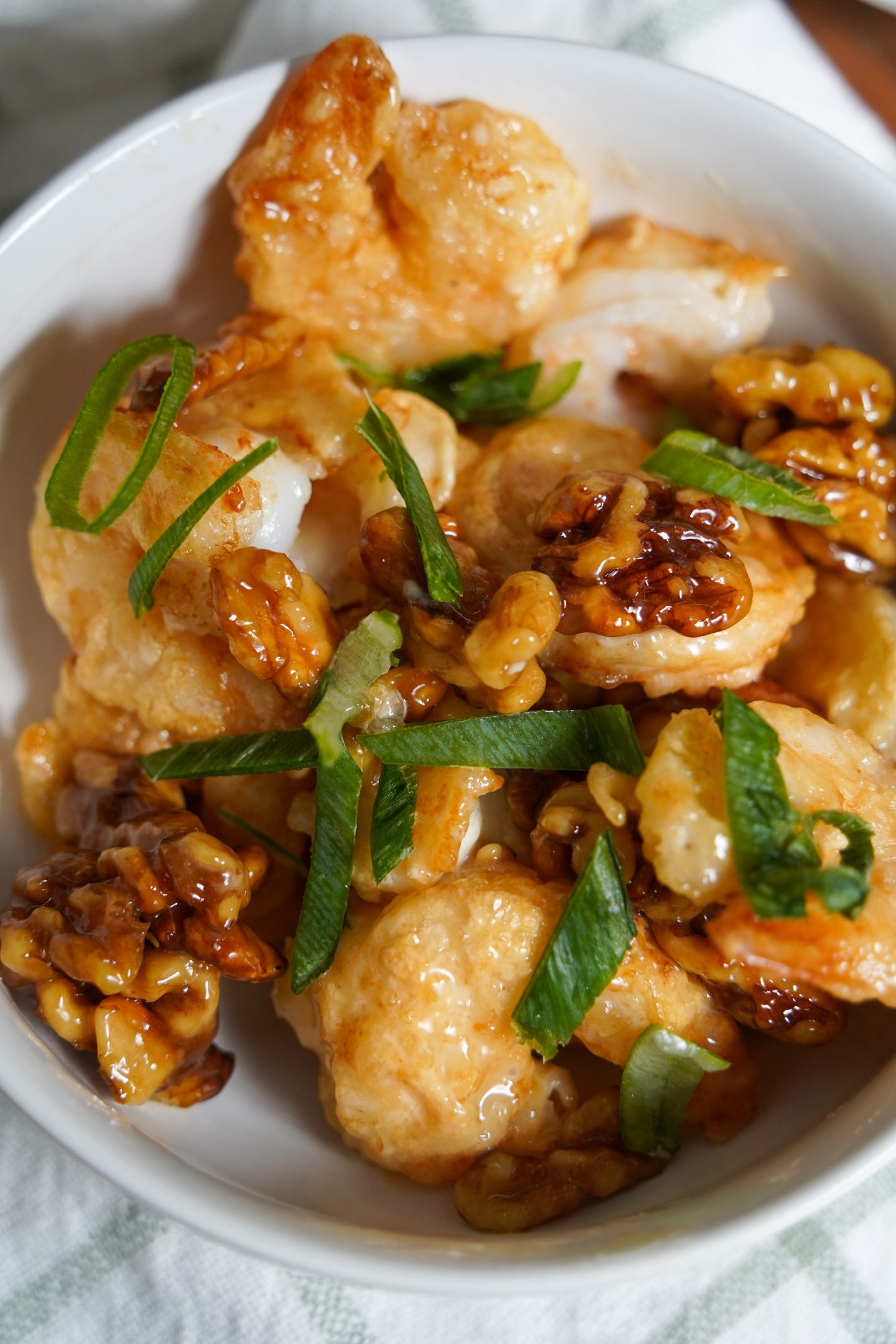 A close-up of homemade Panda Express Walnut Shrimp dupe in a white bowl, showing crispy battered shrimp coated in a honey glaze, garnished with sliced green onions and candied walnuts, placed on a cloth napkin over a wooden surface.