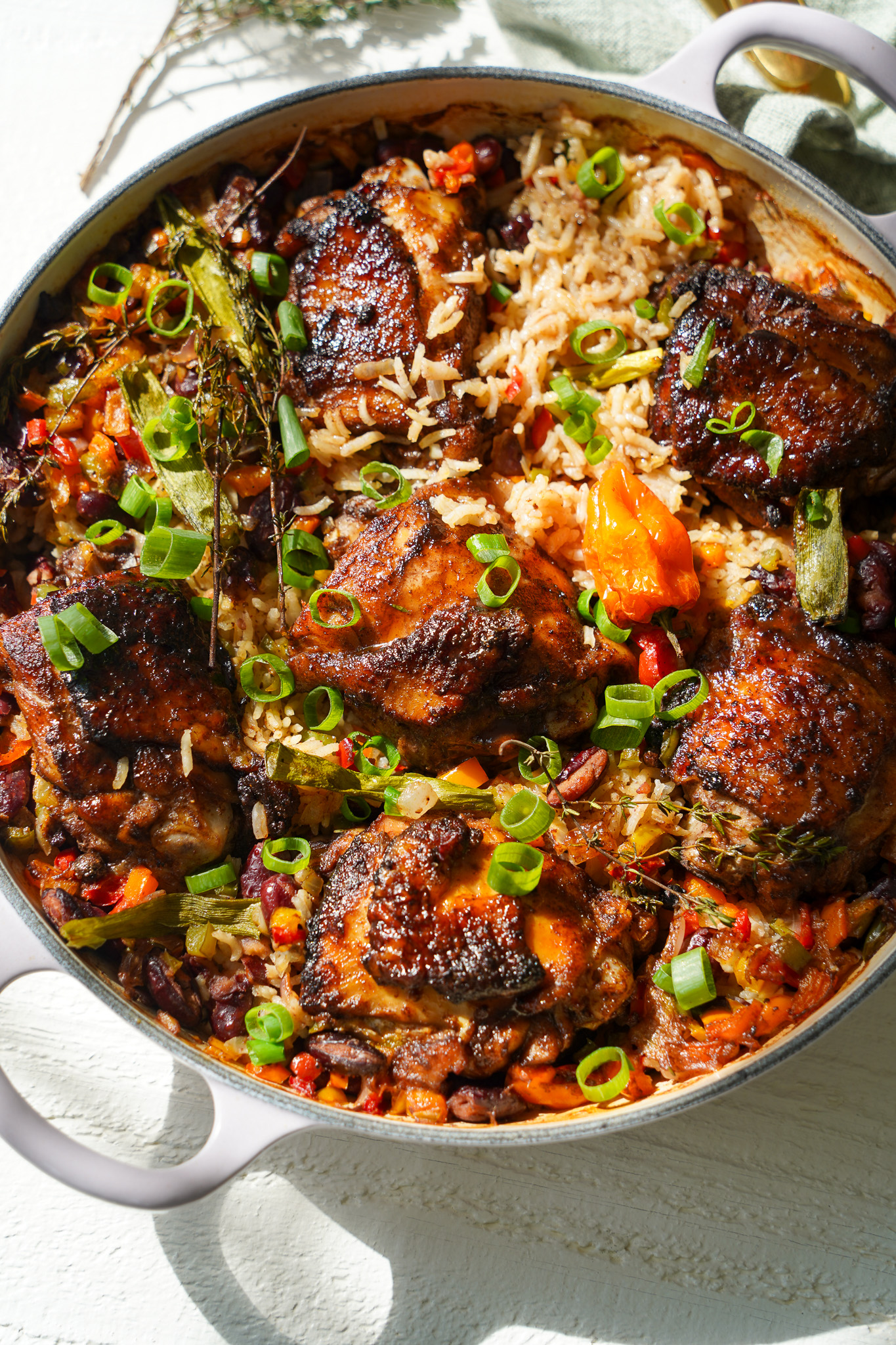 A vibrant, close-up shot of one-pot jerk chicken with rice and peas in a large, round cast iron pan. The dish features golden-brown, spice-rubbed jerk chicken thighs nestled in fluffy, seasoned rice mixed with kidney beans, bell peppers, scallions, and fresh herbs. A whole Scotch bonnet pepper adds a pop of color, while the dish is garnished with chopped green onions and sprigs of thyme. Sunlight highlights the crispy chicken skin and the rich, bold flavors of this Caribbean-inspired meal.