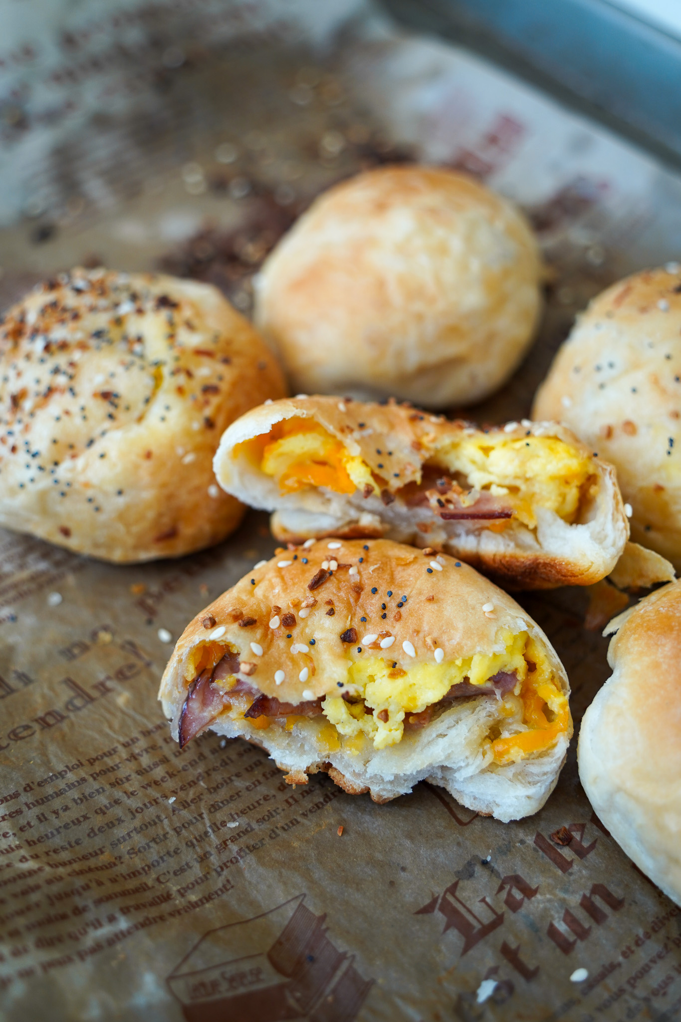 Close-up image of golden baked breakfast biscuits filled with scrambled eggs, cheddar cheese, and slices of ham. The biscuits are topped with Everything But the Bagel seasoning and placed on parchment paper. One biscuit is broken open to reveal the warm, cheesy filling inside.