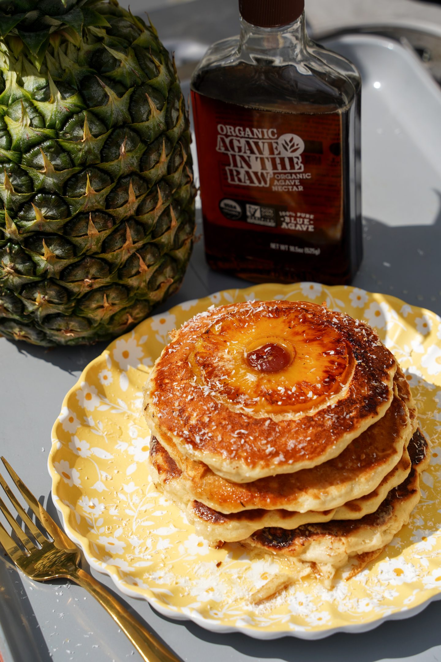 Stack of golden brown pineapple upside down pancakes topped with caramelized pineapple rings and maraschino cherries, served on a yellow floral plate with shredded coconut, next to a bottle of Agave In The Raw and a whole pineapple.