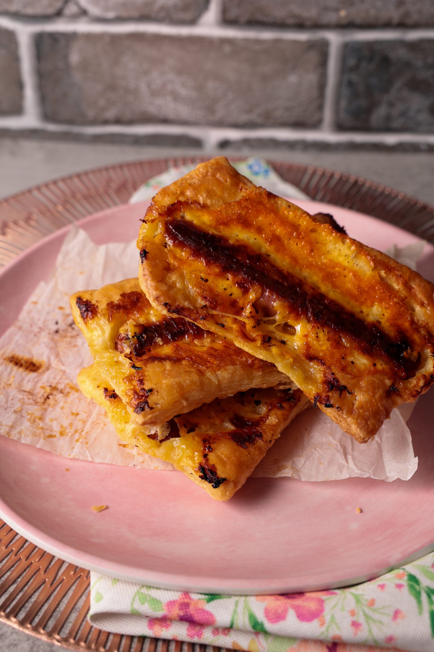 Golden brown upside down hotdog puff pastries stacked on a pink plate with parchment paper, featuring melty cheese and flaky puff pastry edges, perfect for summer BBQs or casual gatherings.