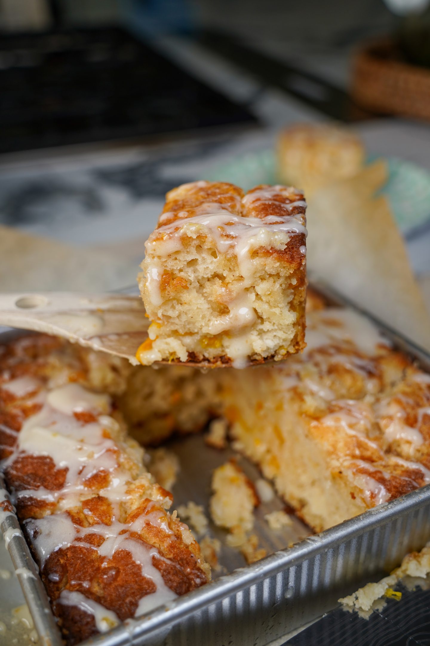 Close-up of a glazed peach butter swim biscuit being lifted from a baking pan, showcasing its fluffy texture, golden edges, and visible peach pieces.