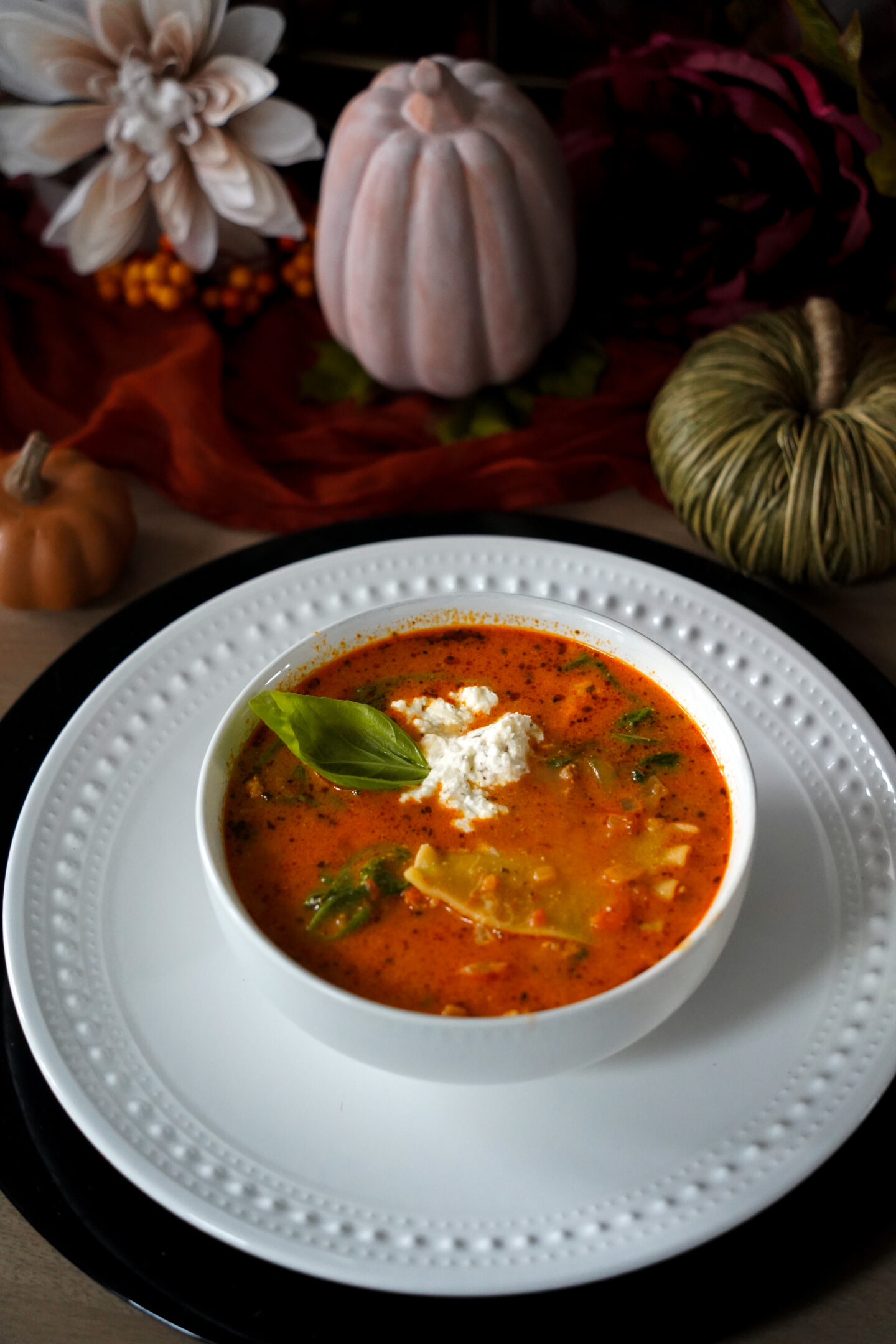 A cozy bowl of lasagna soup topped with ricotta cheese and fresh basil, served in a white bowl on a white plate with fall pumpkin and floral decor in the background.