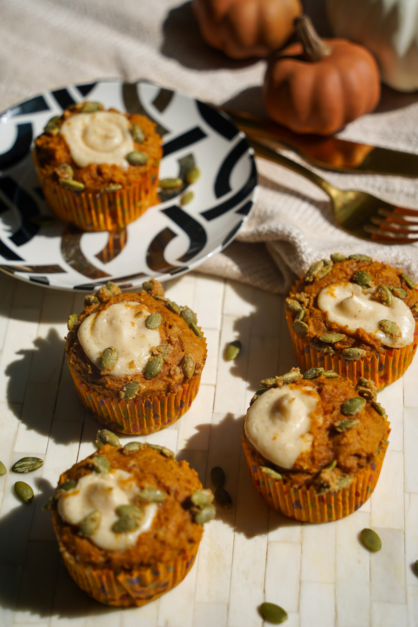 Freshly baked pumpkin cream cheese muffins on a table, topped with candied pumpkin seeds and filled with creamy cheesecake filling, served on a patterned plate with mini pumpkins in the background.
