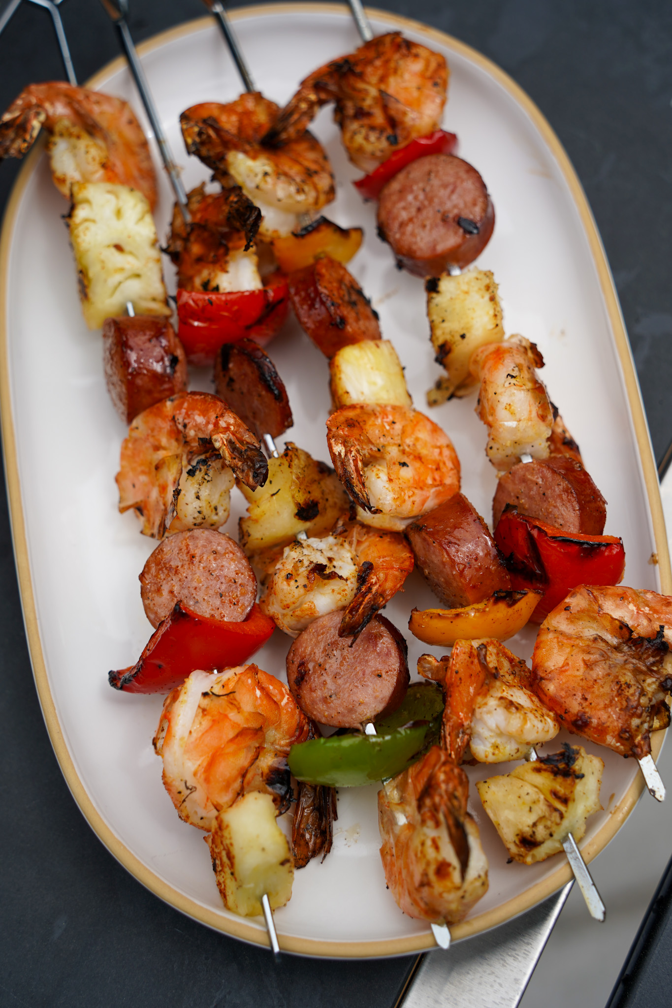 Grilled shrimp, sausage, and pineapple skewers served on a white oval platter, featuring charred shrimp, sliced smoked sausage, caramelized pineapple chunks, and colorful bell peppers with visible grill marks.