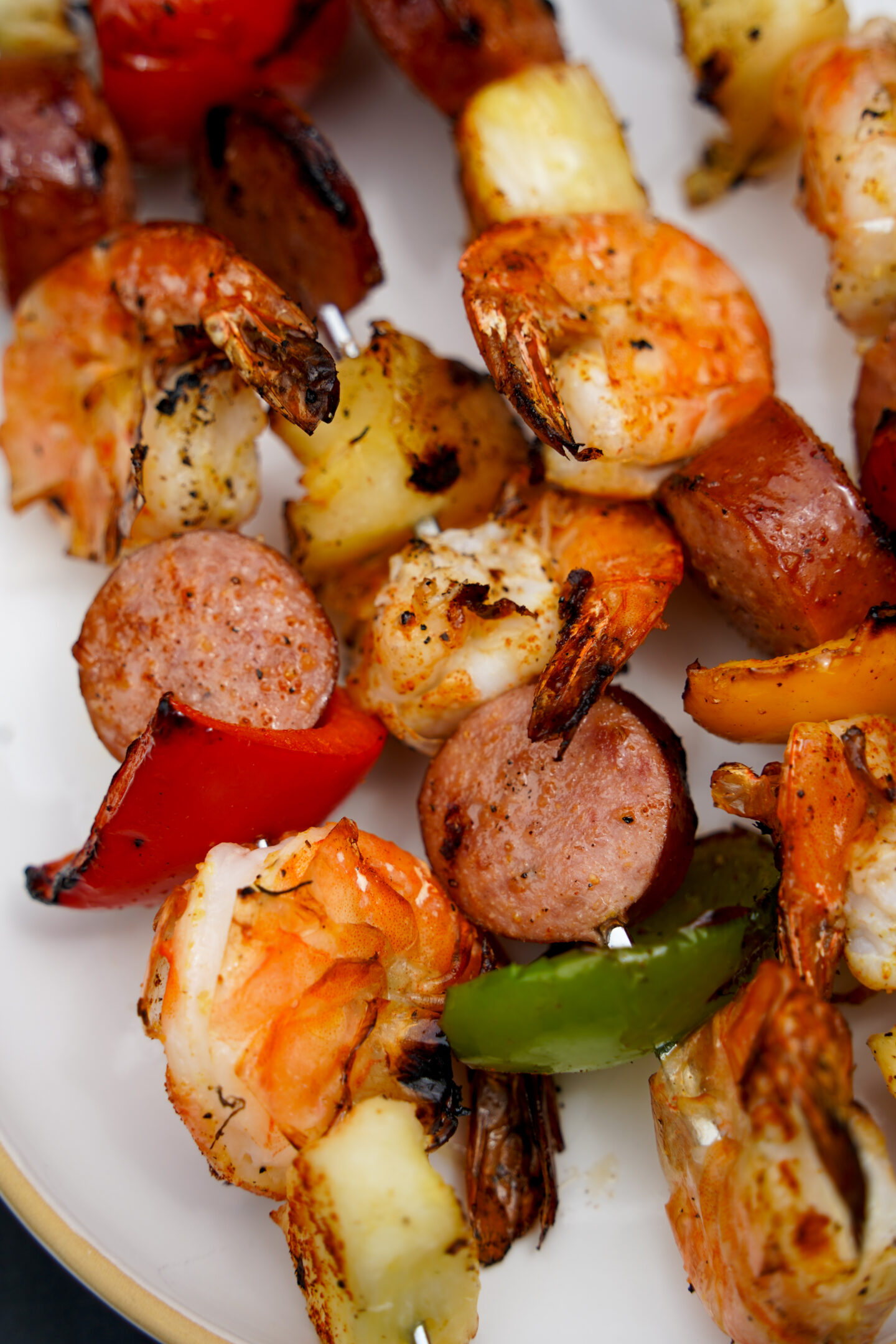 Close-up of grilled shrimp and sausage kebabs layered with charred pineapple chunks and colorful bell peppers on a white serving platter. The skewers feature juicy shrimp with caramelized edges, smoky sausage slices, and vibrant summer vegetables, styled in a bright editorial food photography aesthetic with soft natural lighting and shallow depth of field.