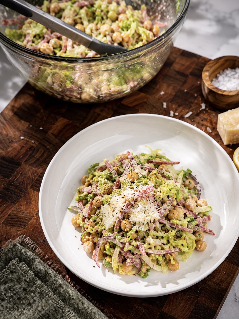 A white ceramic bowl filled with a chopped Italian-style salad sits on a wooden butcher block cutting board in a bright kitchen. The salad features shredded lettuce, chickpeas, red onion, salami, and shaved parmesan tossed in a creamy dressing. In the background, a large glass mixing bowl with extra salad and serving tongs rests near a stovetop, creating a warm, casual food-prep scene.