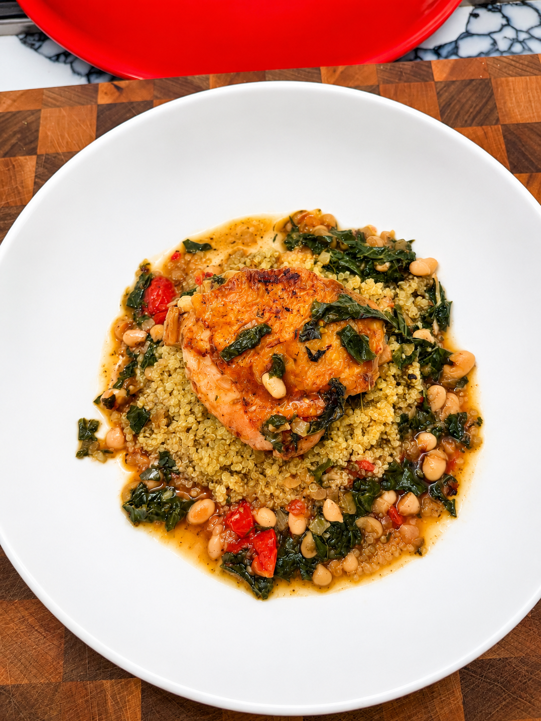 Golden seared chicken thigh served over fluffy quinoa with a Mediterranean-inspired white bean and kale stew in a large white bowl. The dish features tender white beans, wilted kale, diced tomatoes, and a rich savory broth, plated on a wooden butcher block. A wholesome, protein-rich comfort meal with vibrant colors and rustic presentation.